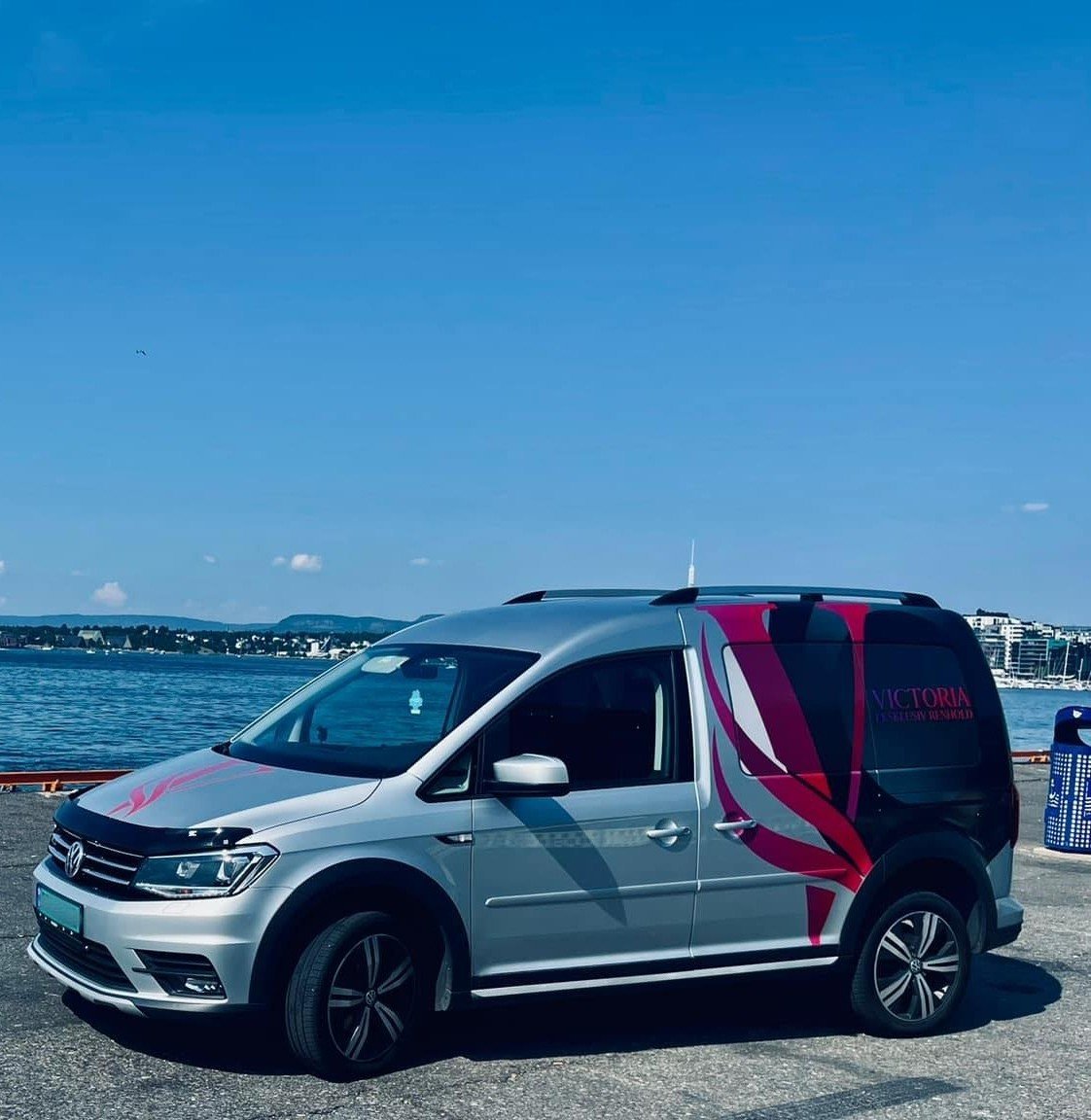 Silver Volkswagen minivan with red and black striped wrap parked at waterfront with blue ocean and mountains in background